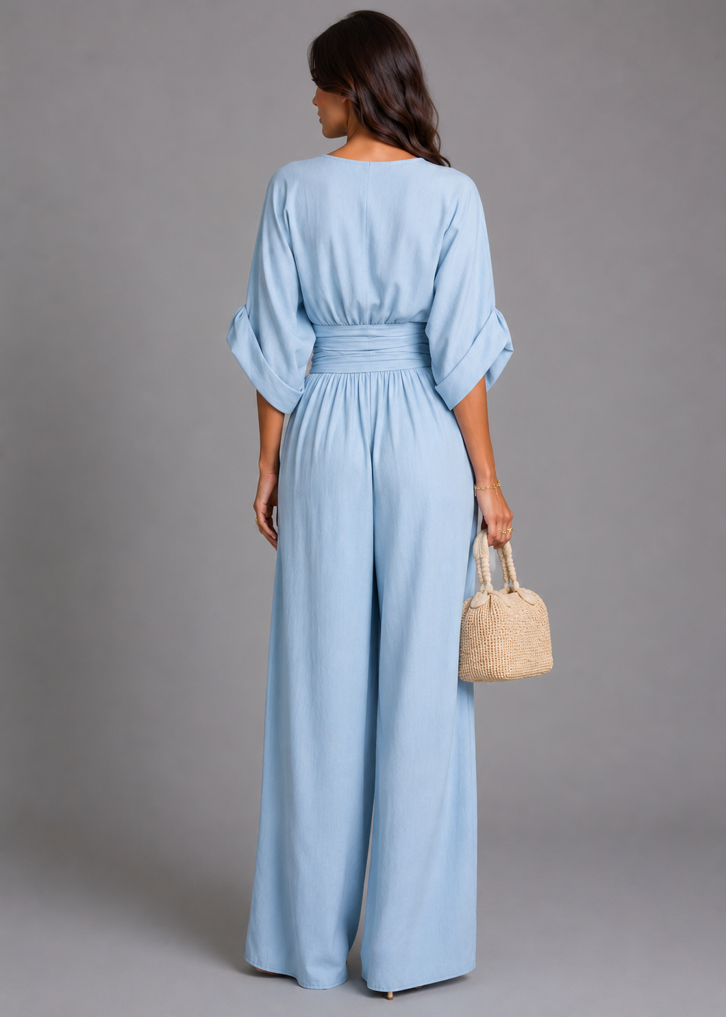 Woman wearing a light blue jumpsuit holding a woven bag against a gray background