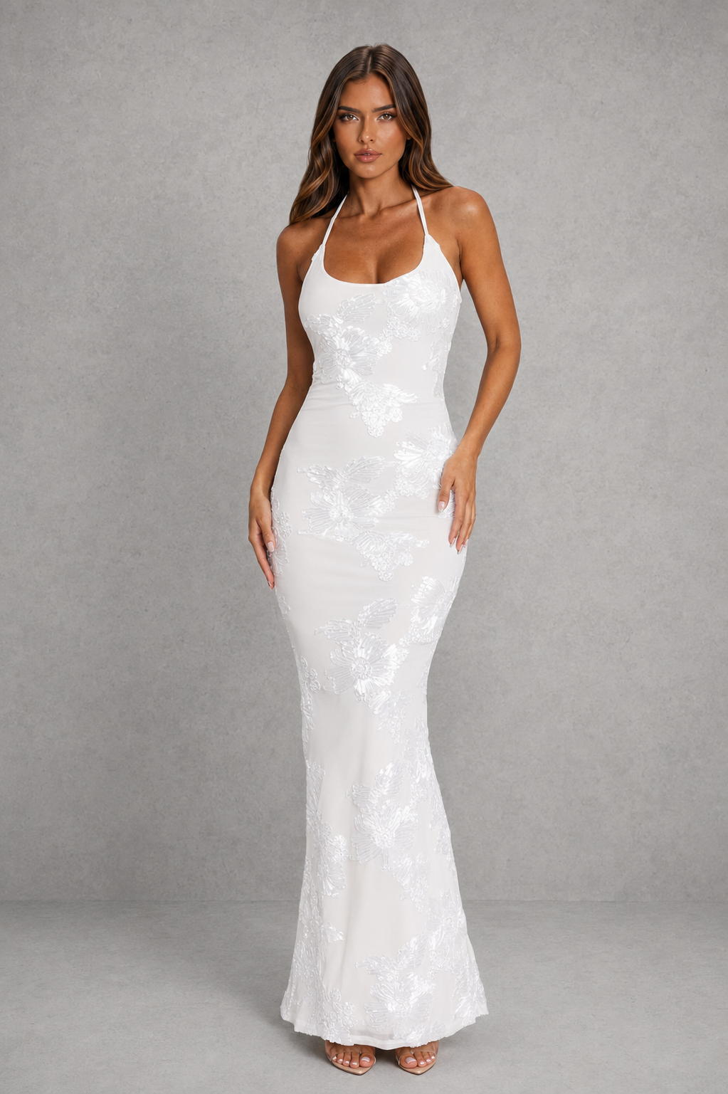 A woman modeling a white sleeveless halter-neck maxi dress with a fitted silhouette and subtle tonal floral lace pattern, set against a neutral gray studio backdrop.