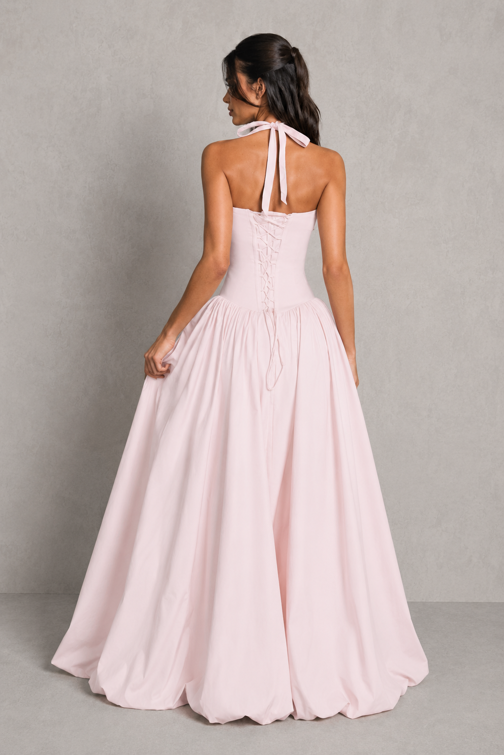 Woman wearing a long, light pink dress with a halter neck design against a plain background.