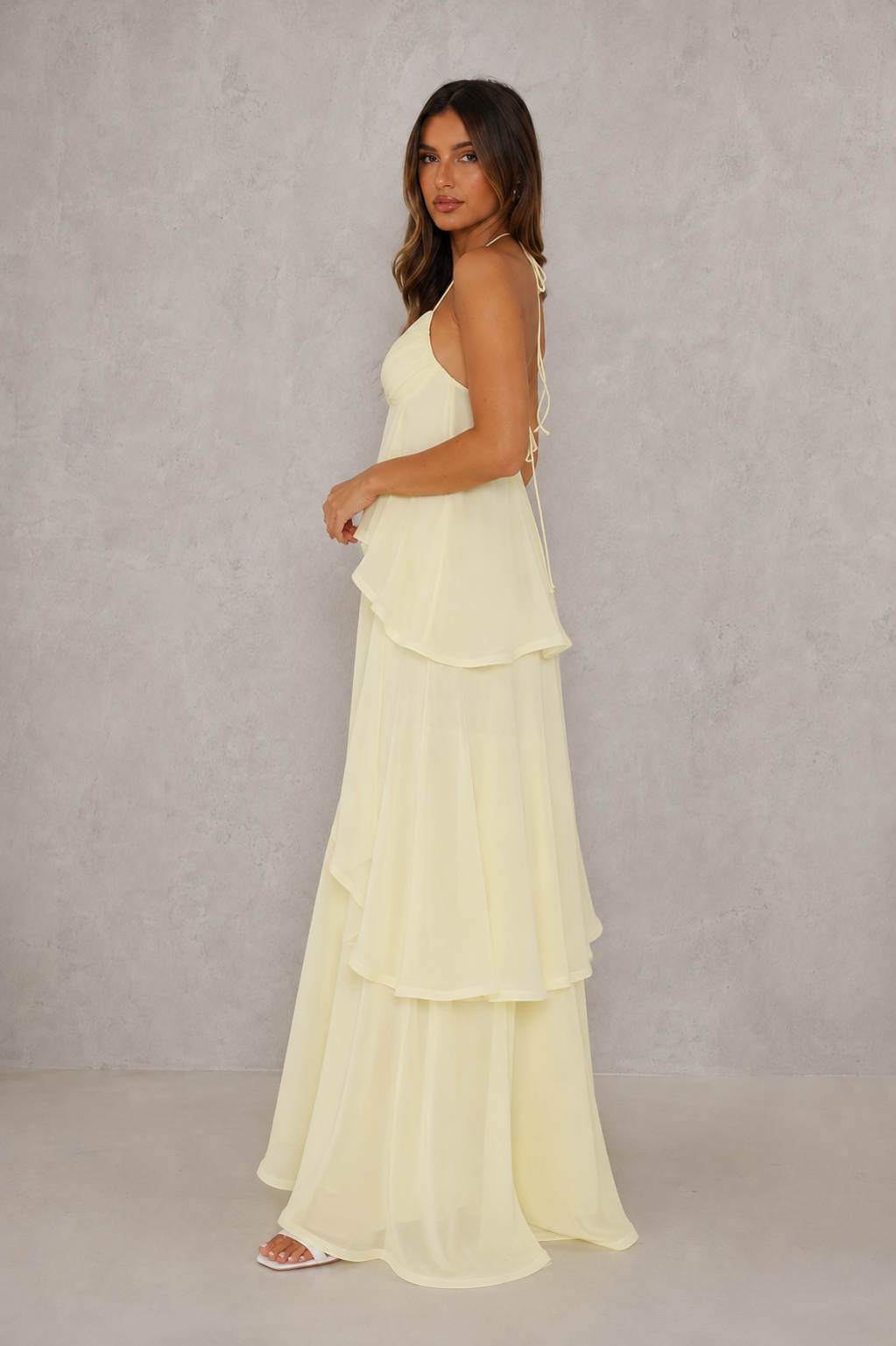 A woman modeling a pale yellow, tiered halter-neck maxi dress against a textured gray studio backdrop.