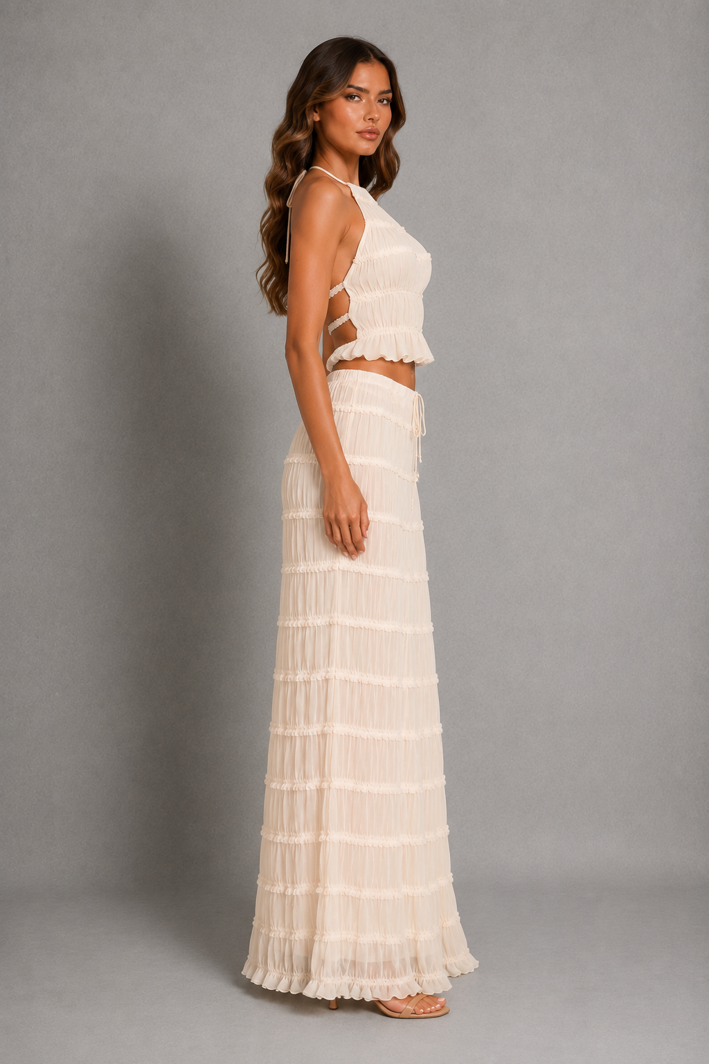 A woman modeling a two-piece cream or off-white two-piece set consisting of a halter crop top with ruffle trim and a matching tiered maxi skirt, set against a plain gray studio background.