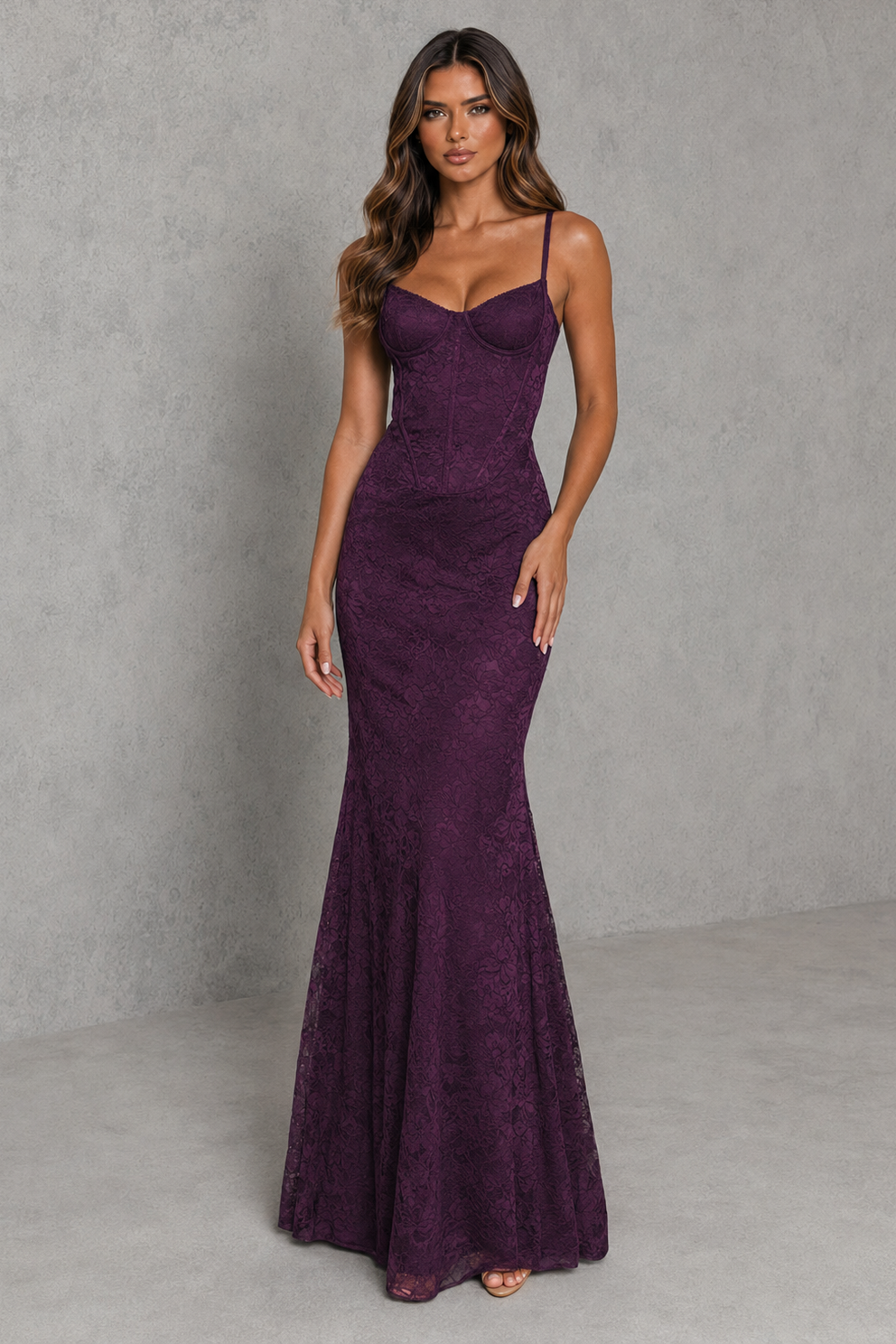 A woman modeling a sleeveless, fitted mermaid-style gown in deep plum purple lace, standing against a textured gray studio backdrop.