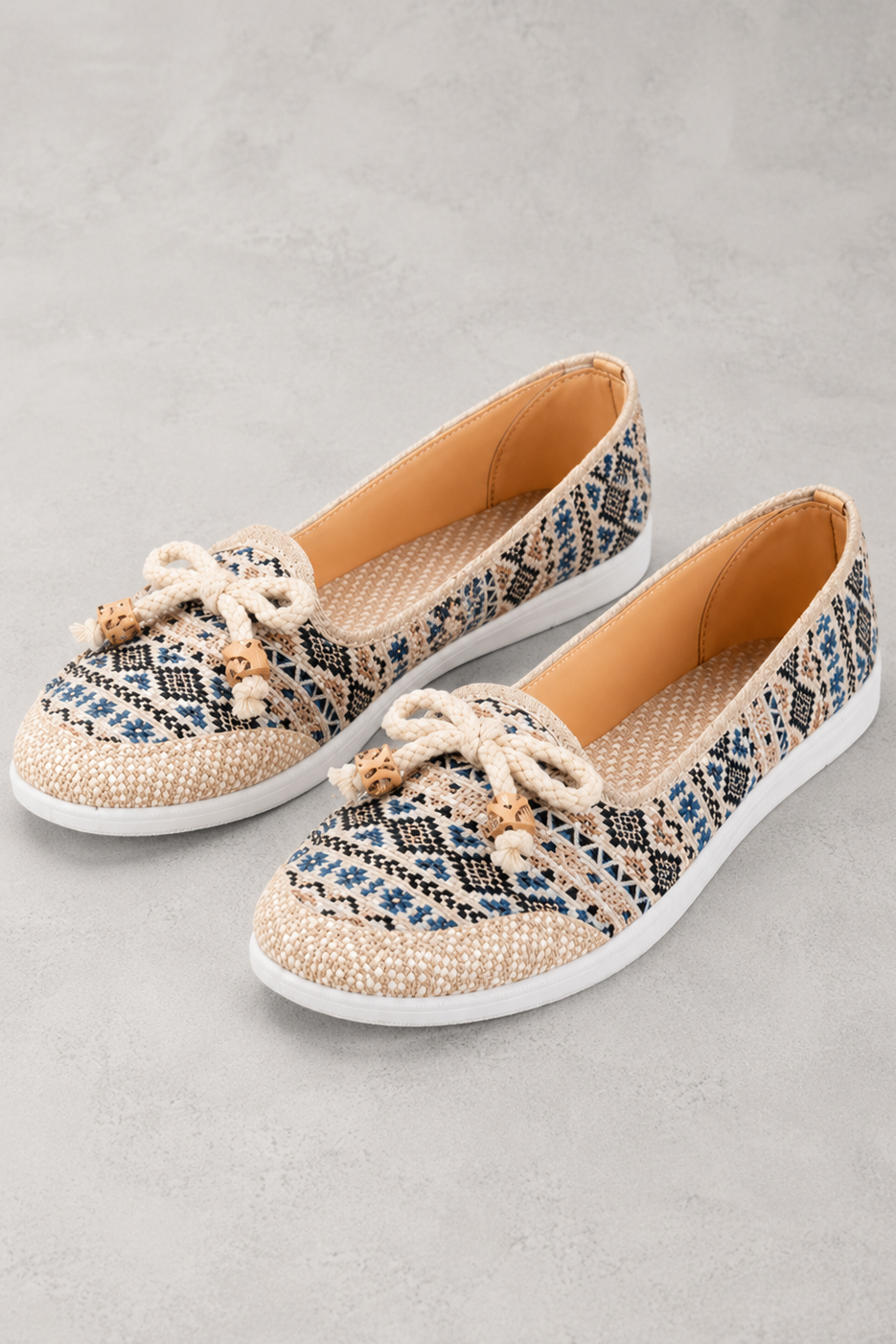 A pair of slip-on espadrille-style flats with a woven snakeskin pattern in beige, blue, and black, featuring cream rope laces with tassel details and white soles, set against a textured gray background.