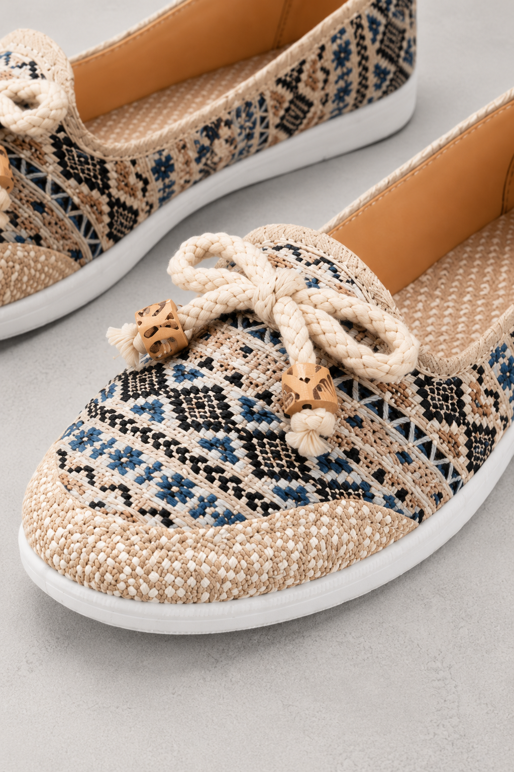 A pair of woven slip-on shoes with a geometric pattern in beige, black, and blue, featuring a thick cream rope bow with wooden beads on the front and a white sole.