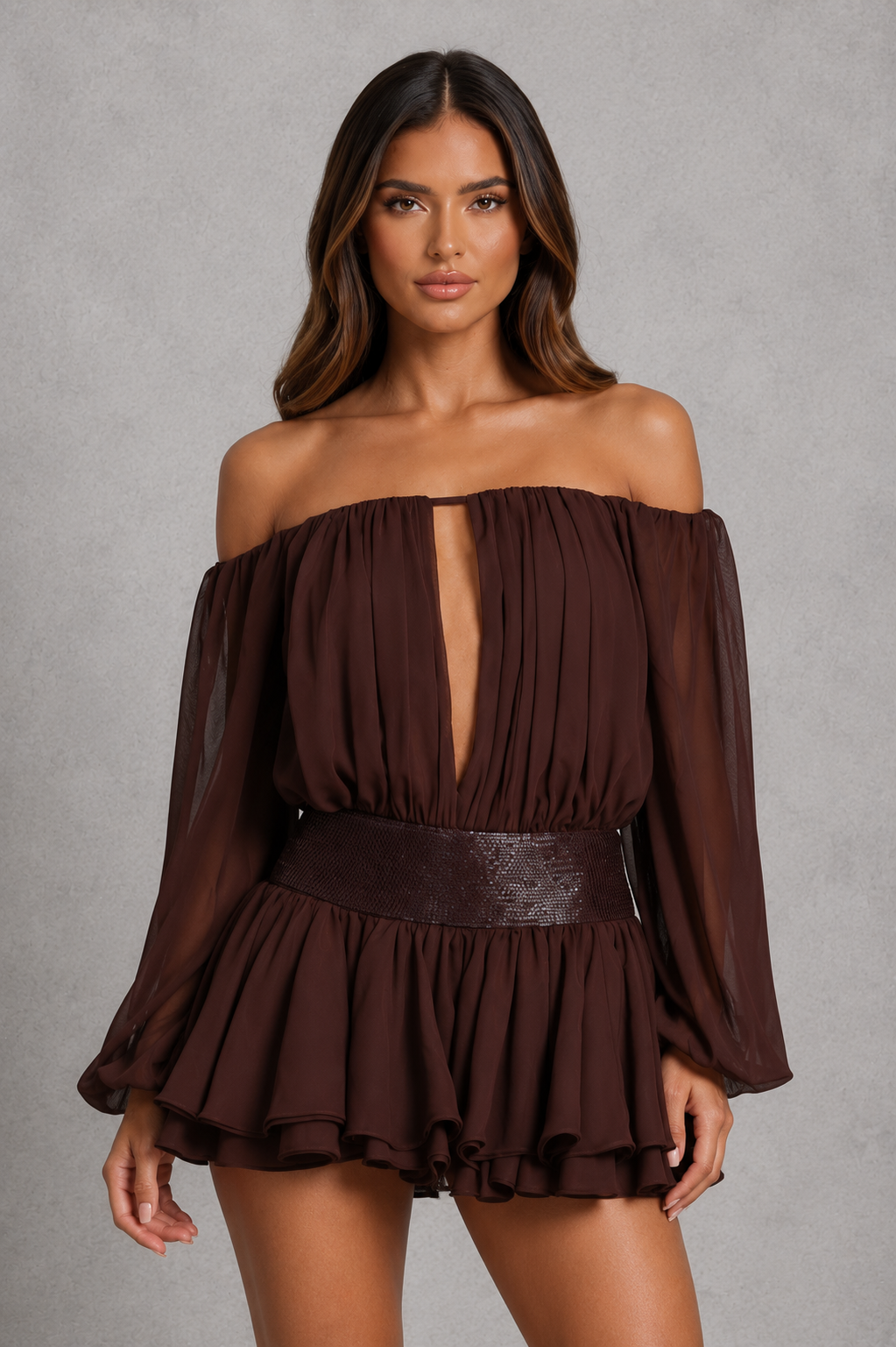 A woman modeling an off-the-shoulder dark brown long-sleeve romper with a deep V-neckline, pleated texture, and a wide matching belt, set against a textured gray studio background.