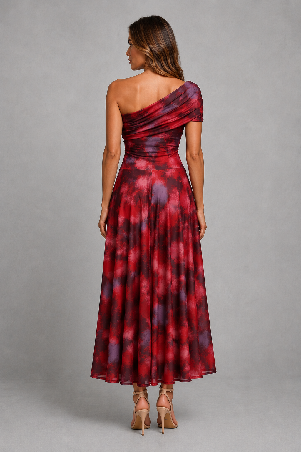 A woman shown from the back wearing a one-shoulder maxi dress with a red, burgundy, and purple tie-dye pattern, paired with beige high-heeled sandals, against a neutral gray studio background.