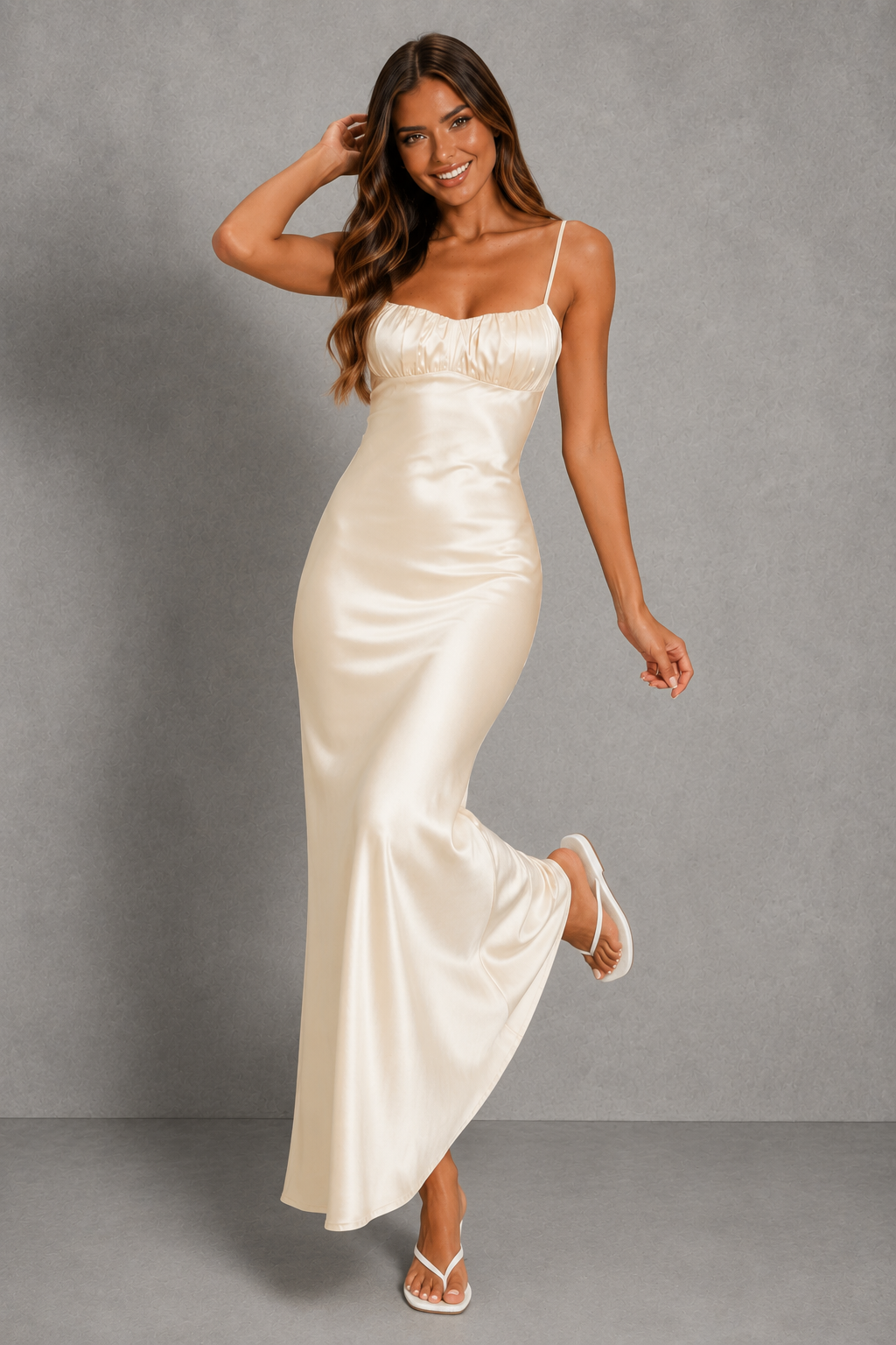 A woman modeling a floor-length satin slip dress in a warm ivory or cream color, standing against a textured gray studio backdrop.