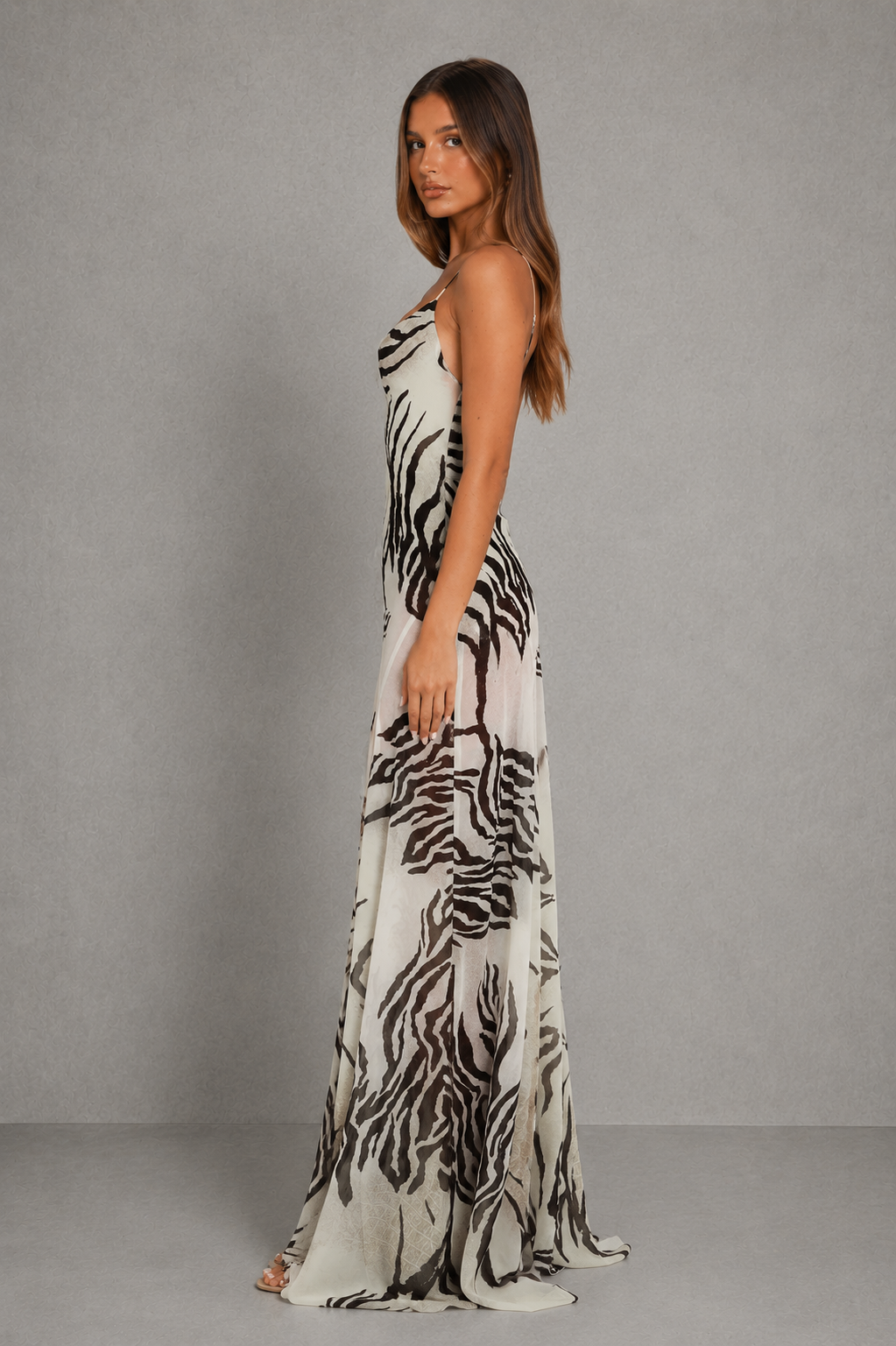 A woman modeling a sleeveless, floor-length maxi dress with a black and white zebra-like animal print, standing against a textured gray studio backdrop.