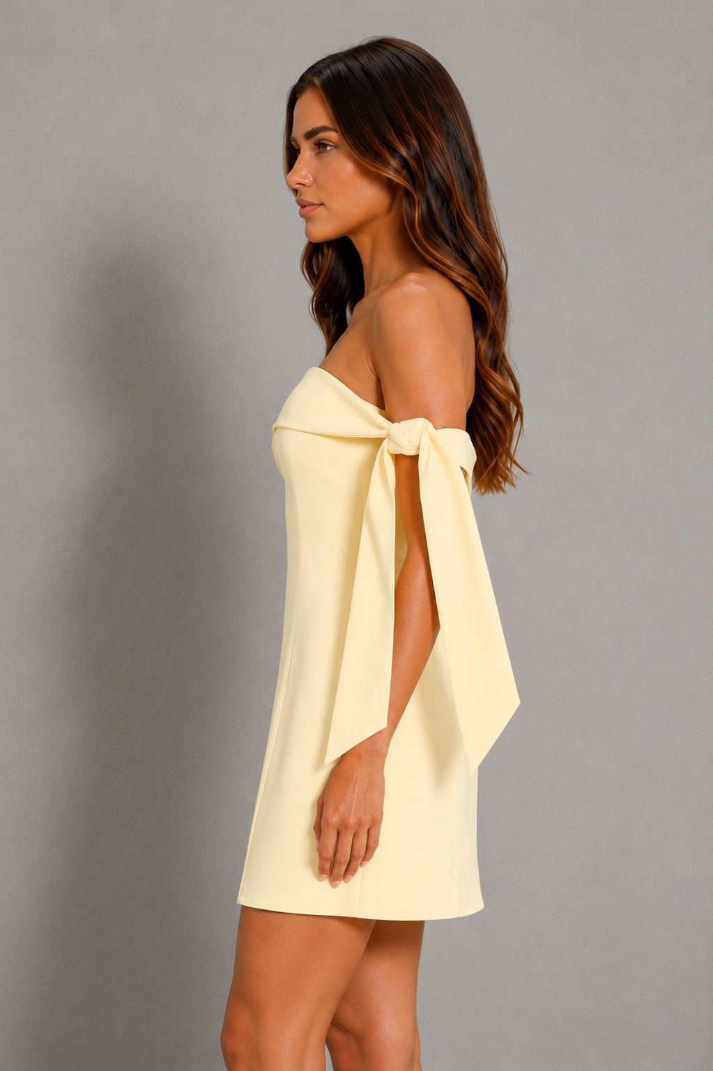 A woman wearing a pale yellow off-the-shoulder mini dress with a large bow detail at the neckline, shown in profile against a plain gray studio background.