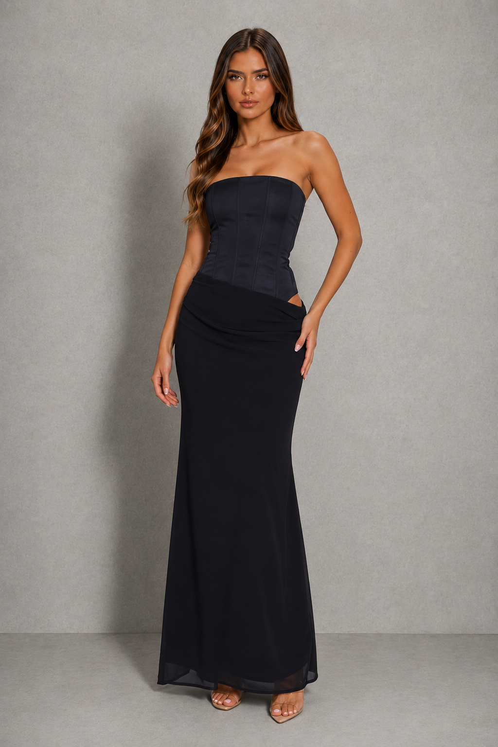 A woman modeling a strapless black maxi dress with a structured corset-style bodice and a flowing skirt, standing against a textured gray studio backdrop.