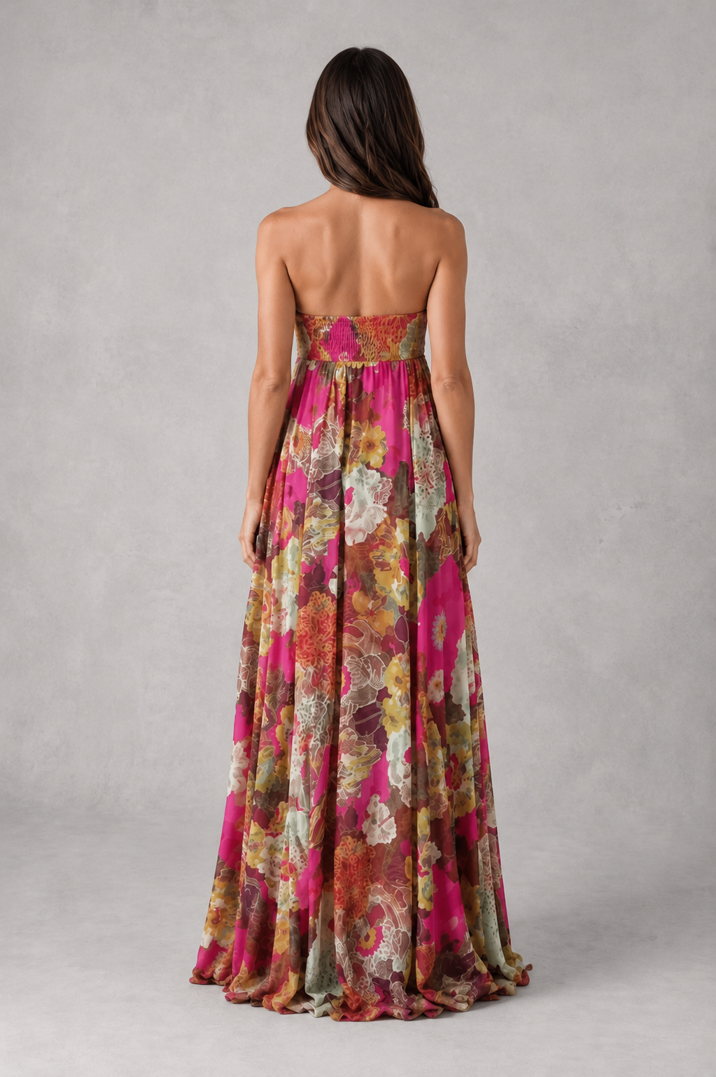 A woman viewed from the back wearing a strapless, floor-length floral gown with a smocked bodice, set against a neutral gray studio background.