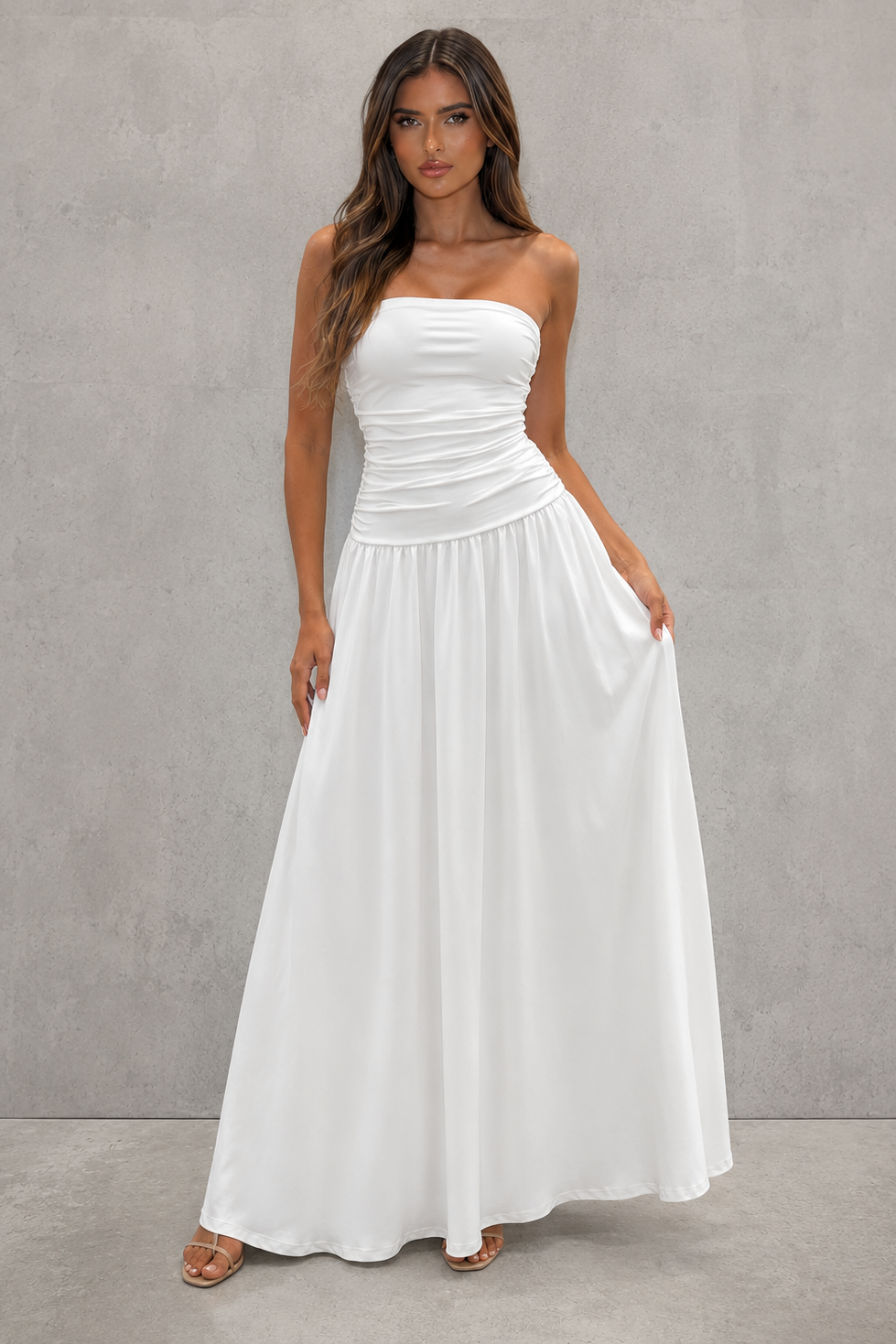 A woman modeling a strapless white maxi dress with a ruched bodice and flowing skirt, standing against a textured gray studio wall.