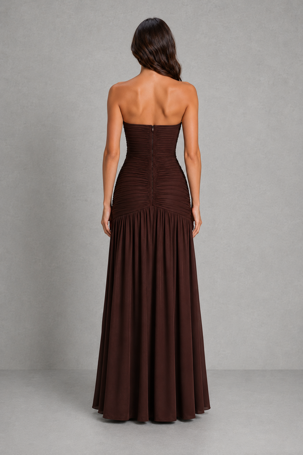 A woman shown from the back wearing a strapless floor-length gown in a very dark brown or near-black color, standing against a plain gray studio backdrop.