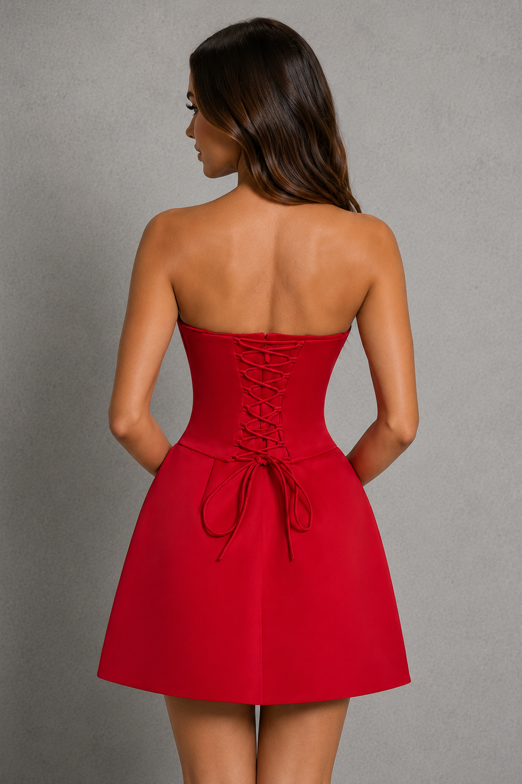A woman shown from the back wearing a bright red strapless mini dress with a corset-style waist and lace-up back, photographed against a plain gray studio background.