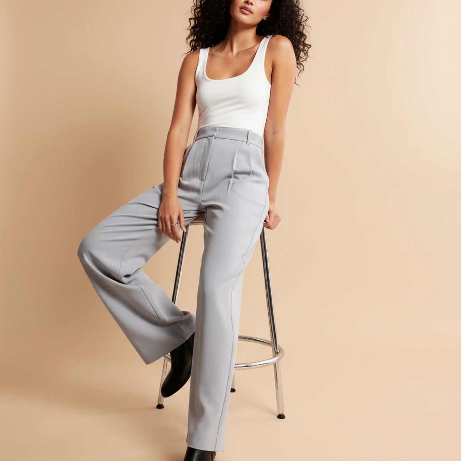 Woman wearing a white tank top and gray high-waisted pants sitting on a stool against a beige background