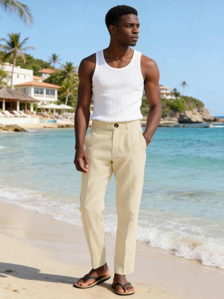 Man in white tank top and beige pants standing on a beach with ocean and palm trees in the background