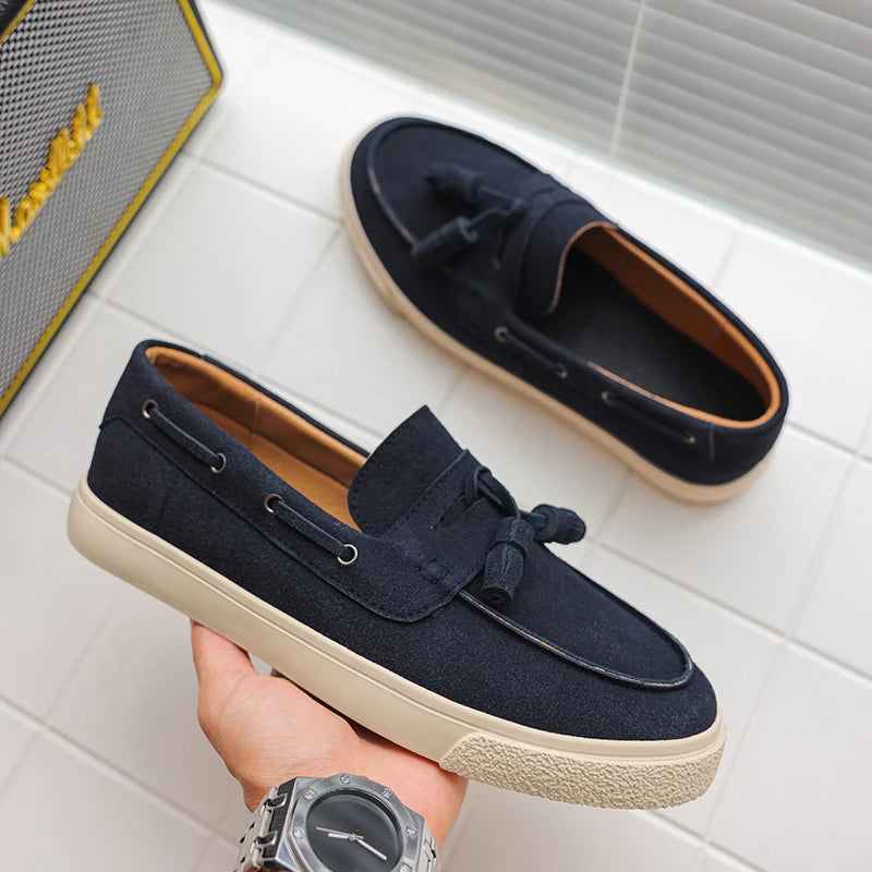 Navy loafers with a beige sole held by a hand on a light gray tiled floor.
