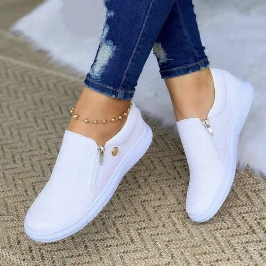 White slip-on sneakers worn with blue jeans on a textured surface.