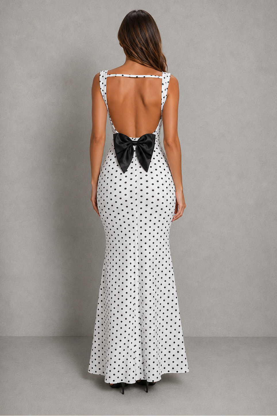 A woman viewed from the back wearing a white sleeveless maxi dress with a black polka dot pattern and a large black bow at the open back, standing against a plain gray studio backdrop.