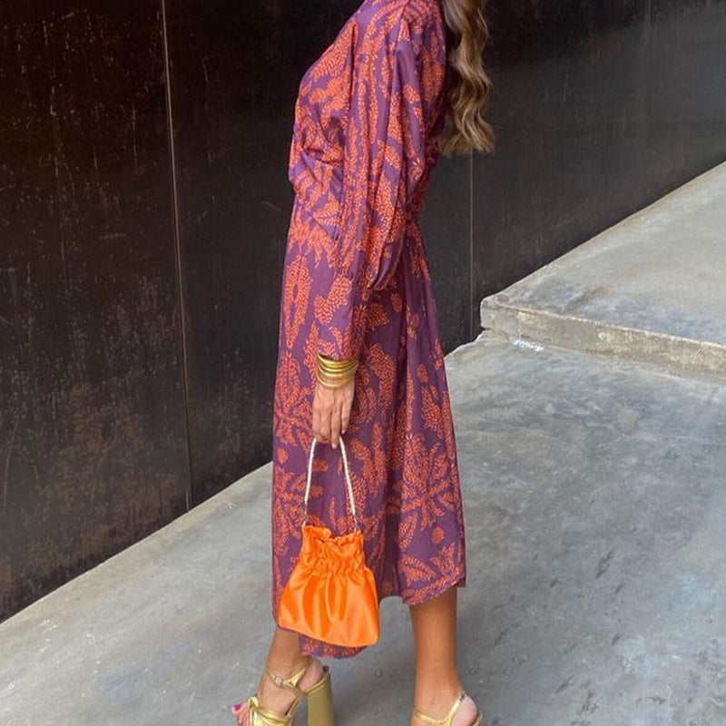 Woman in a patterned dress holding an orange clutch against a black wall.