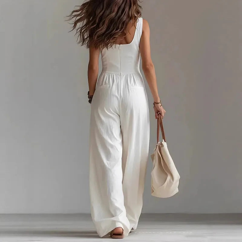 Woman wearing a white sleeveless jumpsuit holding a beige bag against a plain background