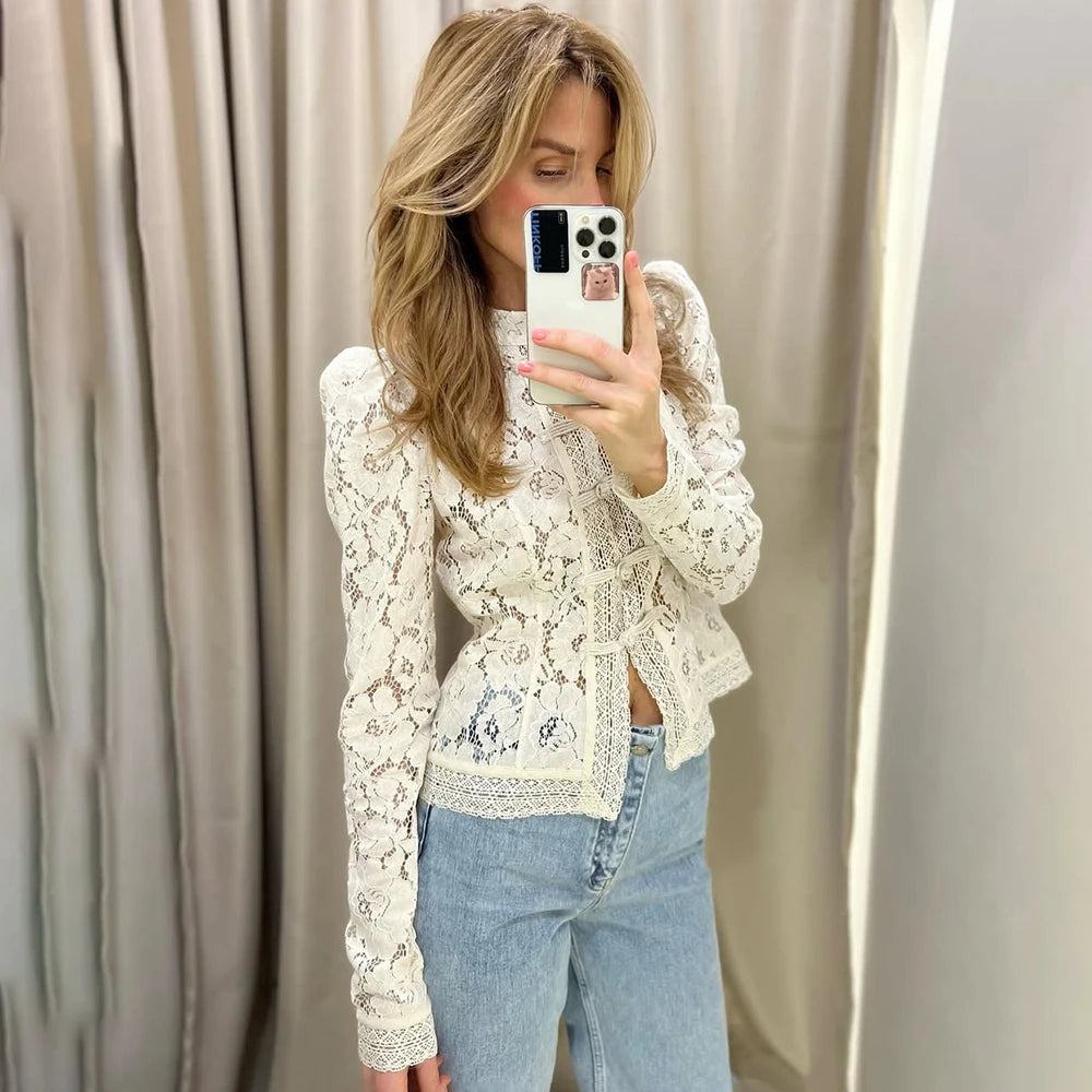 Woman taking a mirror selfie wearing a white lace top and blue jeans against a beige curtain.
