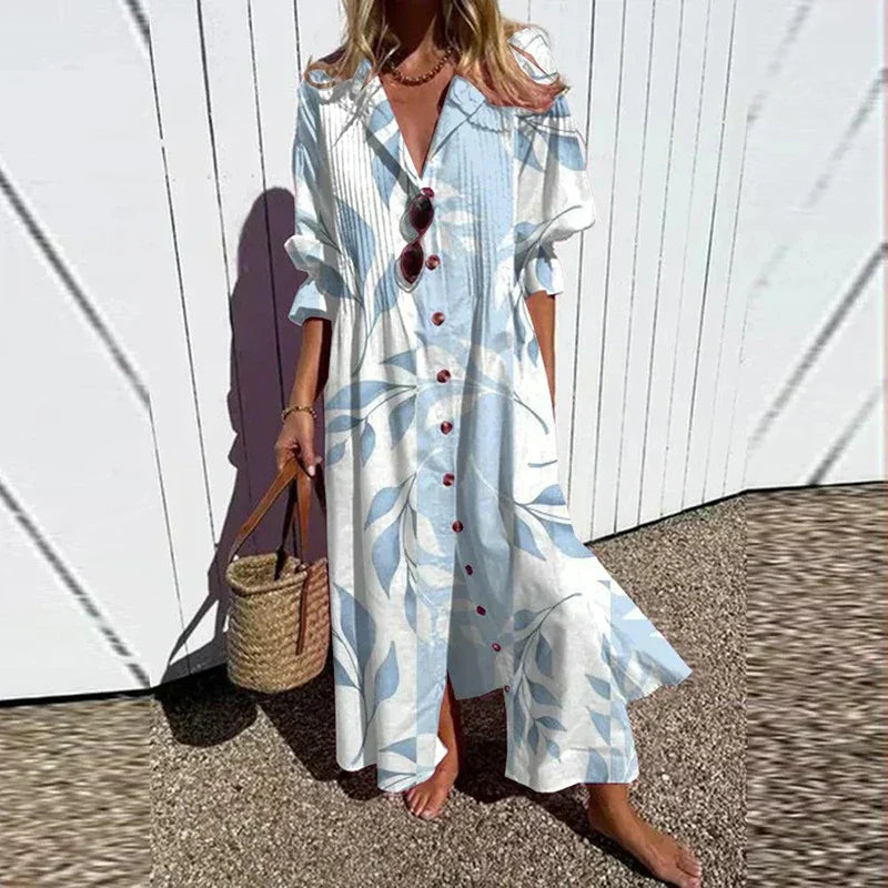 Woman wearing a long, light blue dress with white patterns, standing against a white wall.