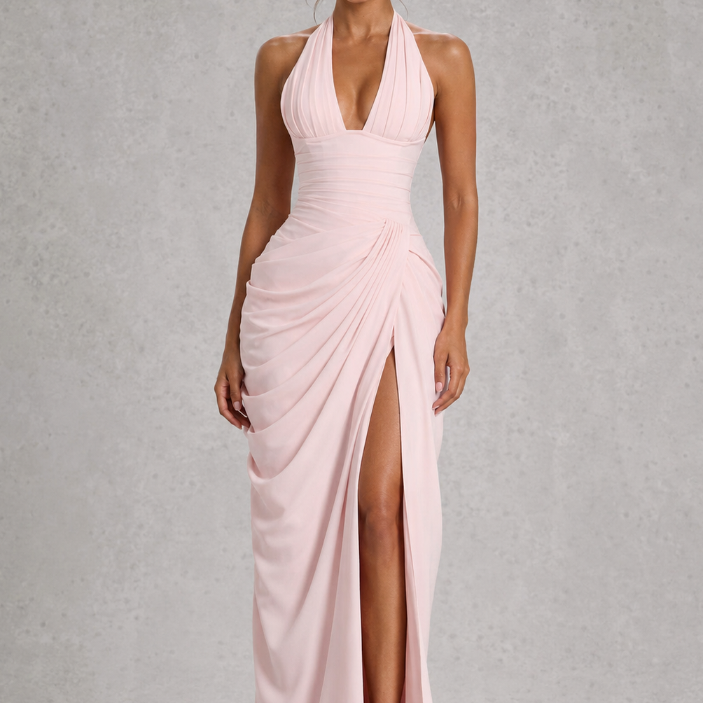 A woman modeling a light pink halter-neck maxi dress with a draped, gathered skirt and a high thigh slit, set against a textured gray studio background.