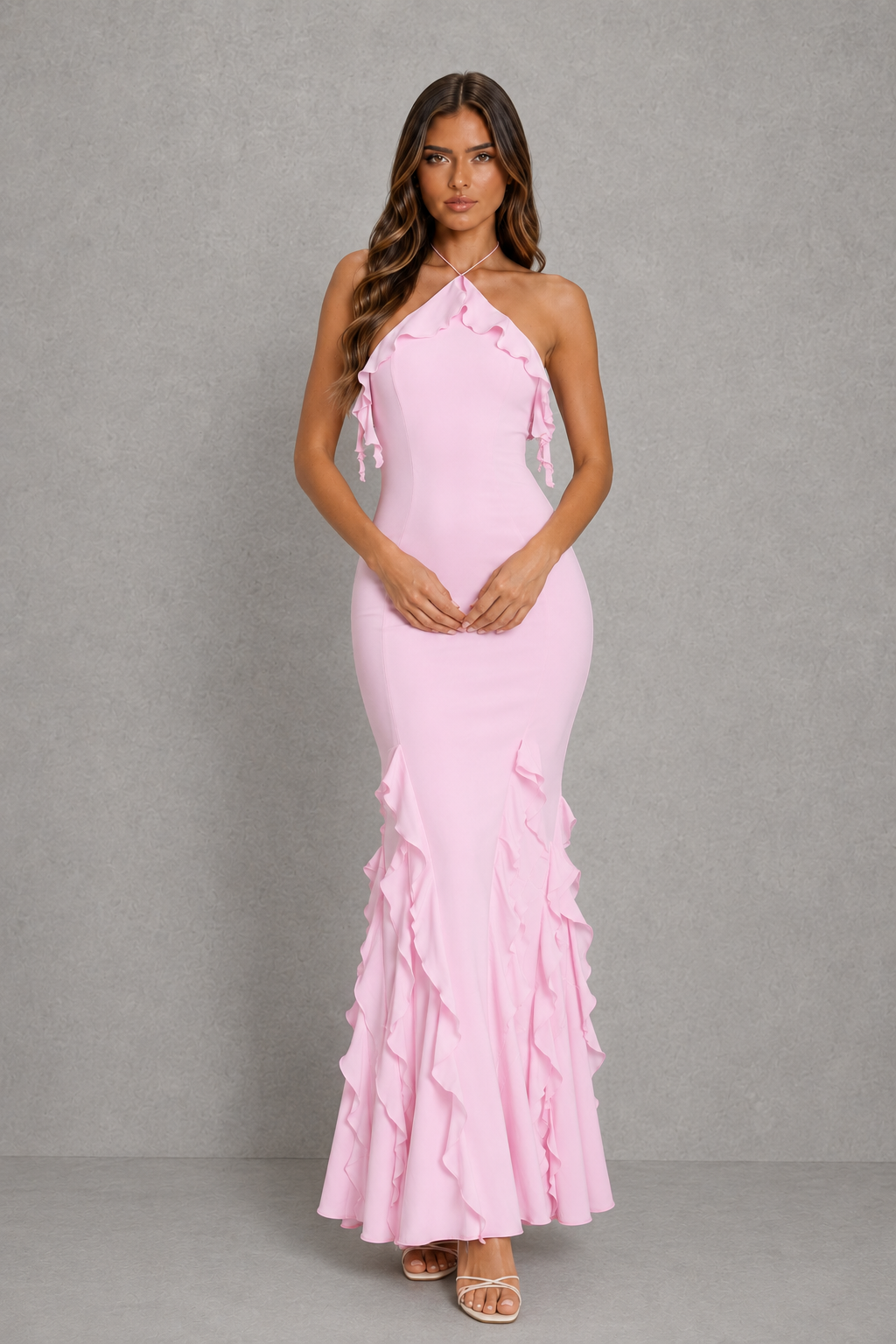 A woman modeling a light pink halter-neck maxi dress with ruffle details along the neckline and cascading down the skirt, set against a textured gray studio background.