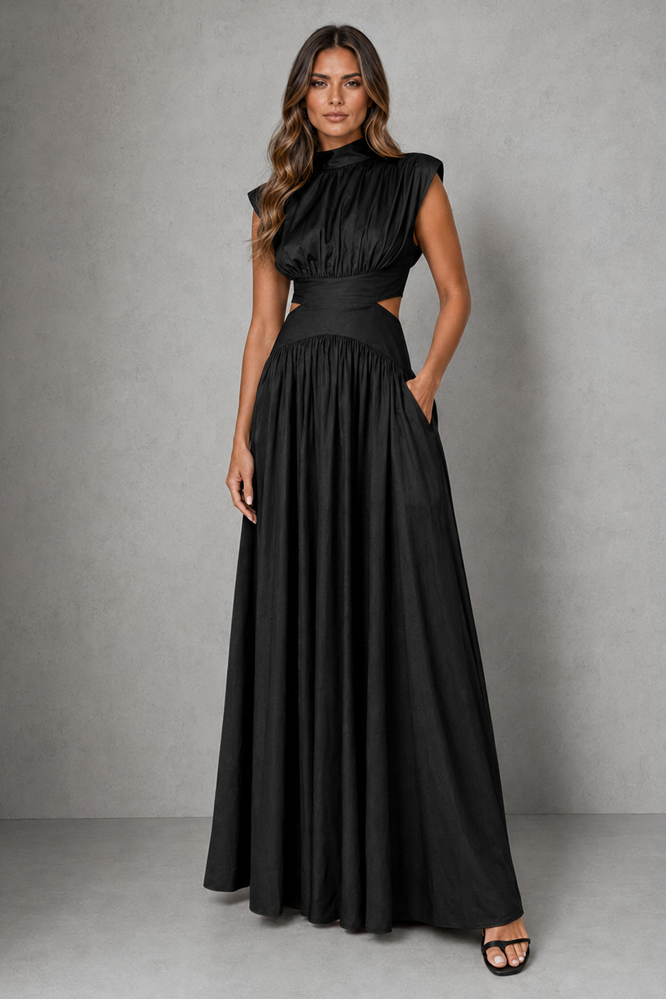 A woman modeling a floor-length black dress with a high mock neck, cap sleeves, side cutouts at the waist, and a flowing skirt, standing against a textured gray studio backdrop.
