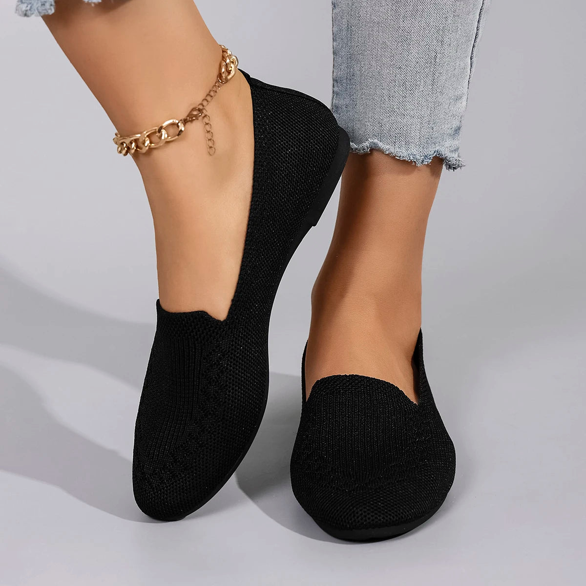 Black slip-on shoes worn with gold anklets on a neutral background
