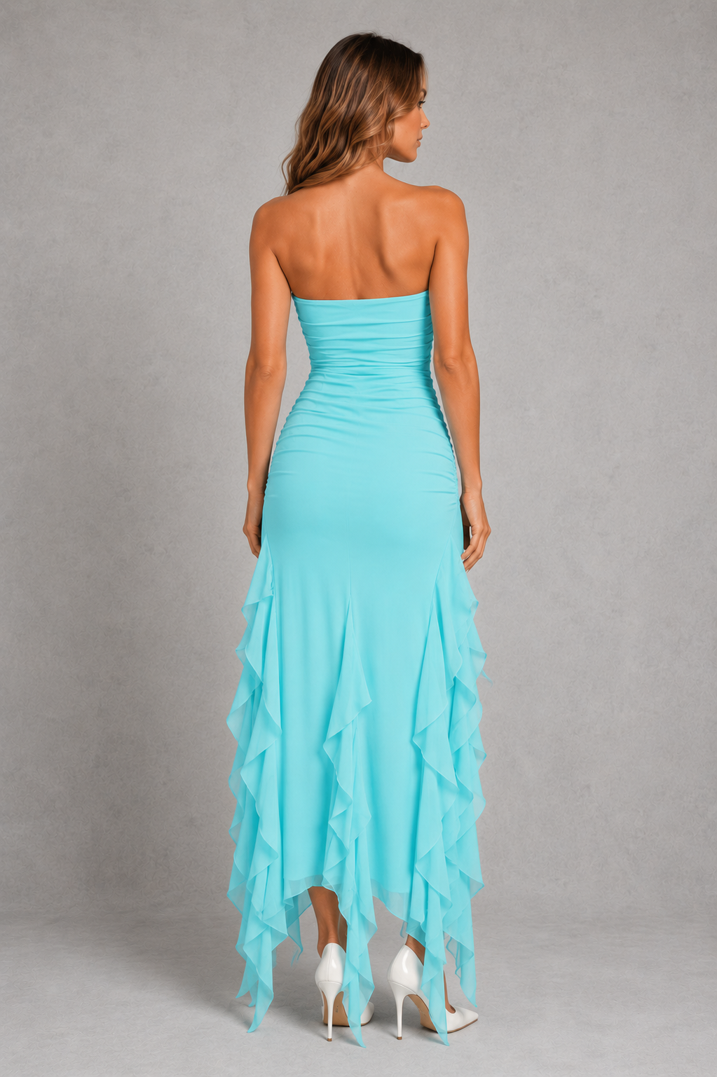 A woman modeling a strapless turquoise blue maxi dress from the back, featuring a ruched bodice and layered ruffle hem, set against a neutral gray studio background.