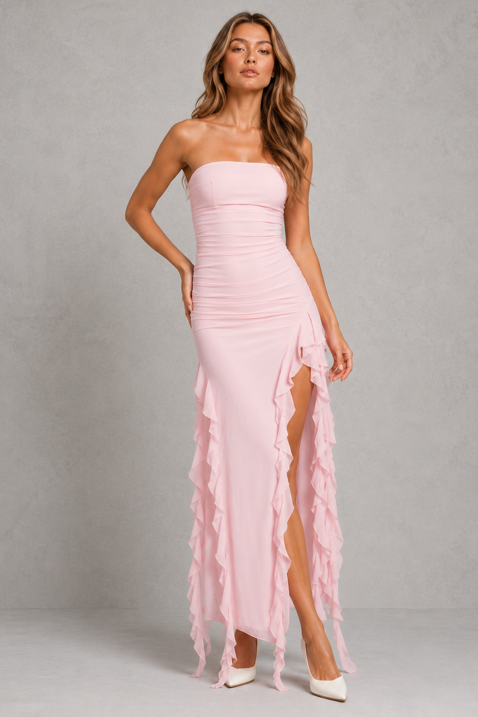 A woman models a strapless light pink maxi dress with a ruched bodice, high slit, and cascading ruffle hem against a textured gray studio backdrop.
