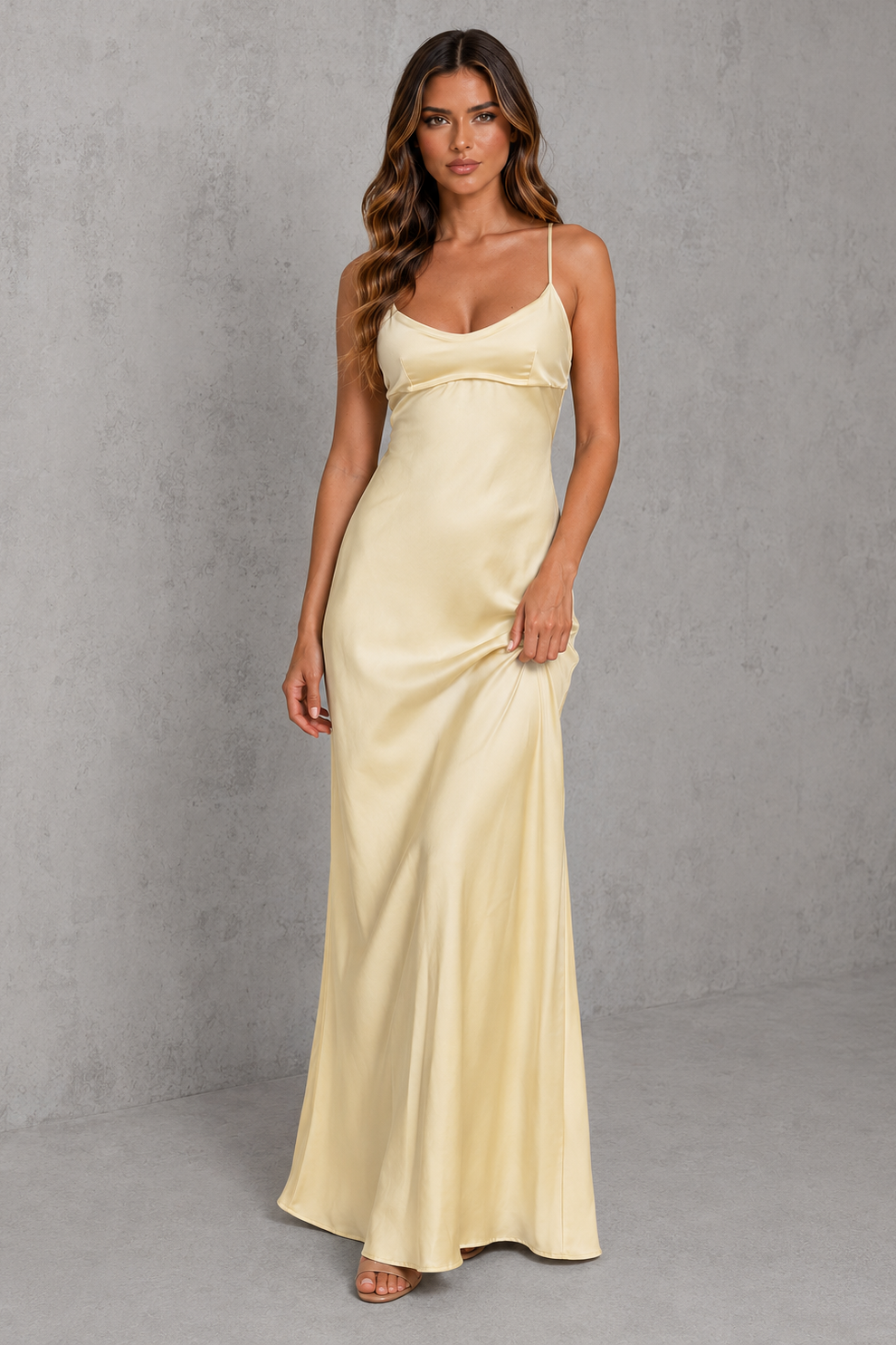 A woman modeling a floor-length satin slip dress in a pale cream or light beige color, standing against a textured gray studio backdrop.