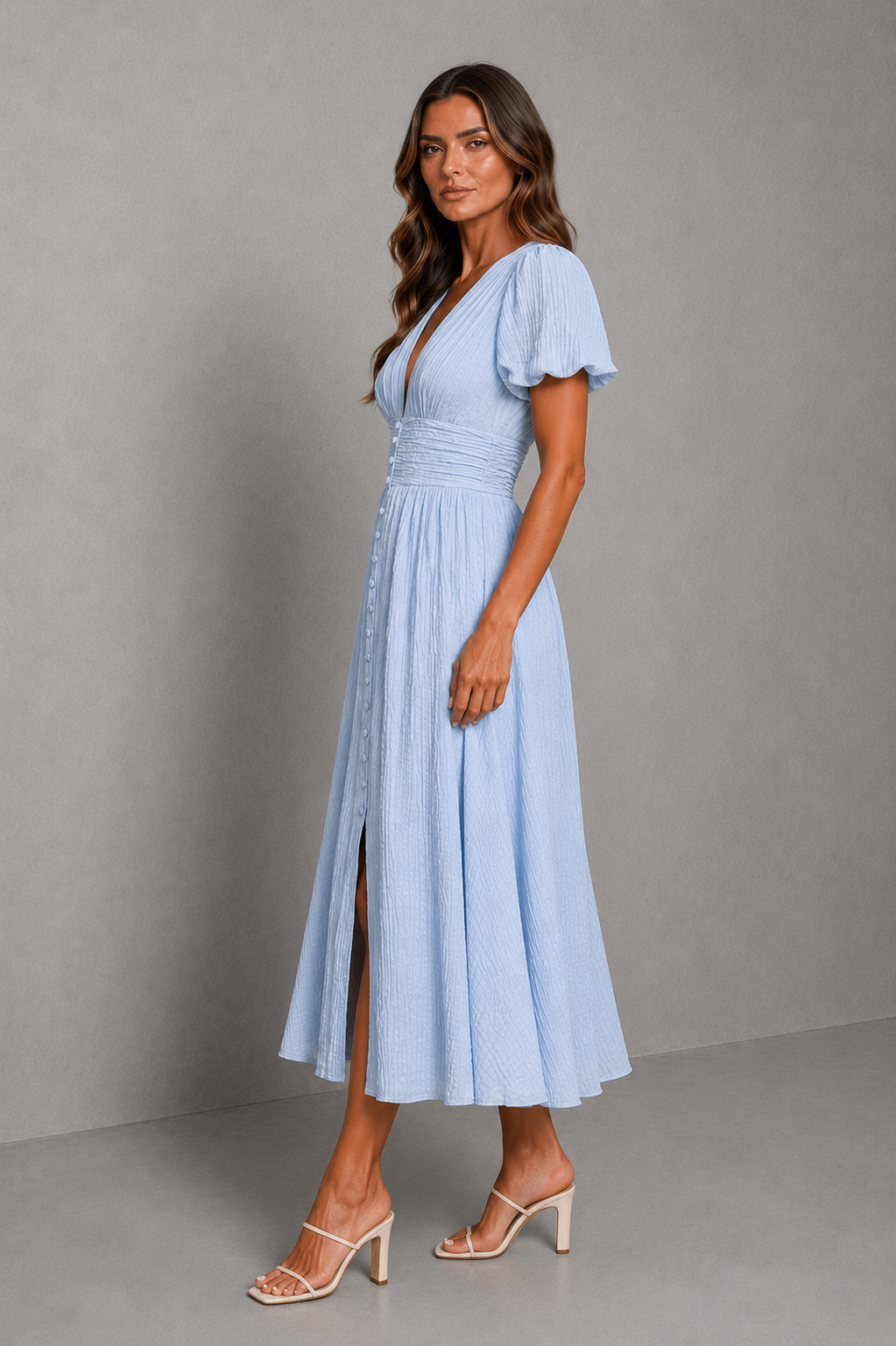 A woman modeling a light blue midi dress with a wrap-style V-neckline, short flutter sleeves, a gathered waist, and a front slit, paired with beige strappy high-heeled sandals against a gray studio backdrop.