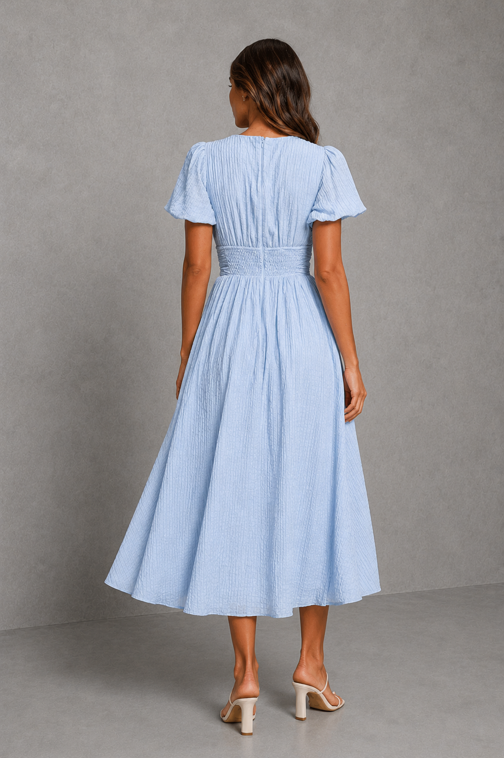 A woman viewed from the back wearing a light blue midi dress with short flutter sleeves, a smocked waist, and a flared skirt, paired with beige heeled sandals against a gray studio backdrop.