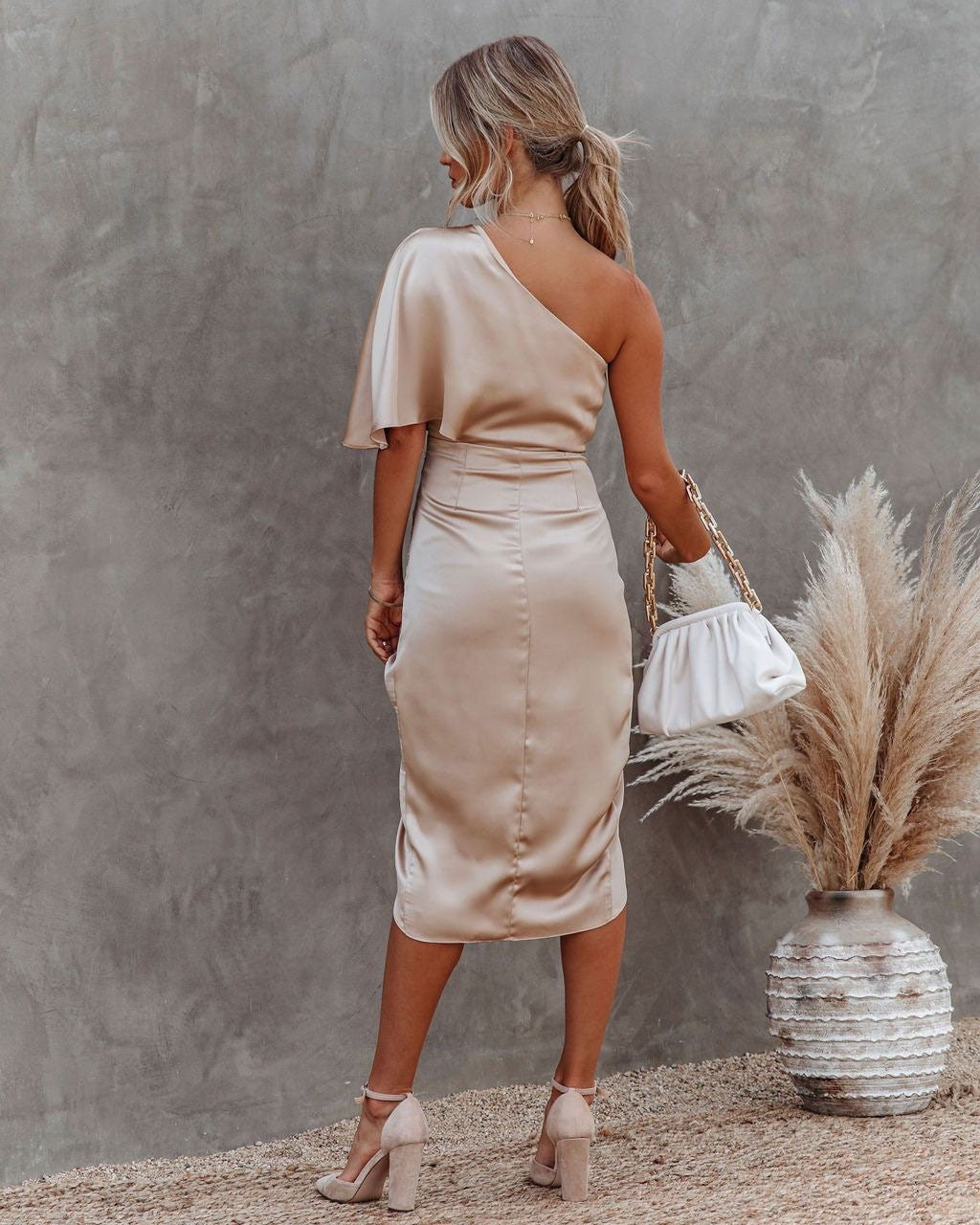Woman in a beige one-shoulder dress standing against a gray wall with dried pampas grass and a vase.