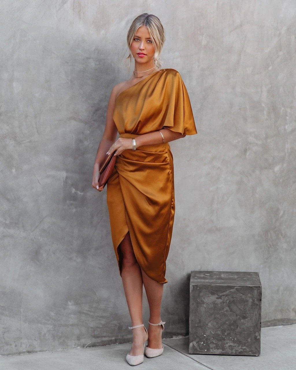 Woman wearing a gold one-shoulder dress against a gray concrete wall.