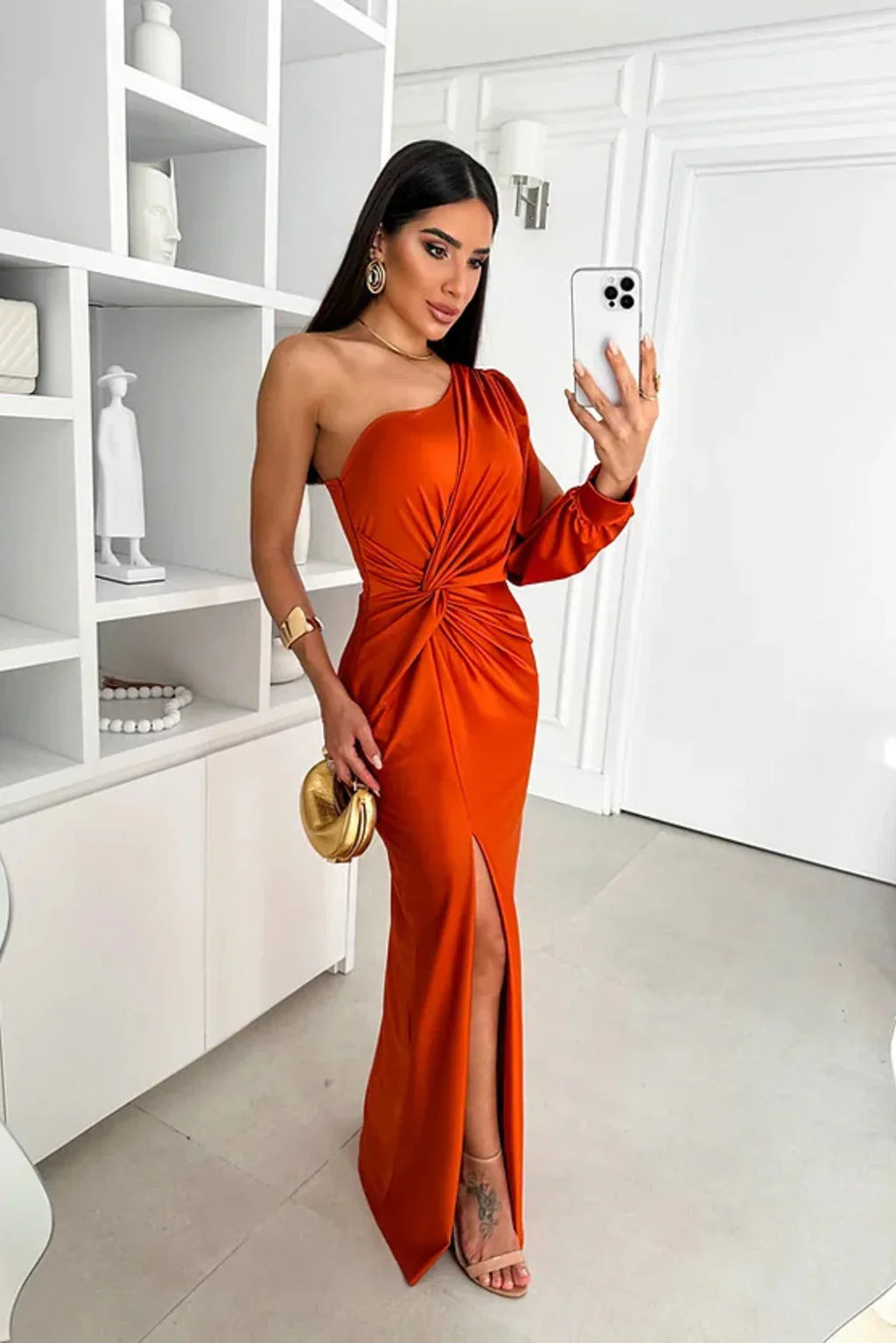 Woman in an orange one-shoulder dress taking a selfie in a modern interior setting.