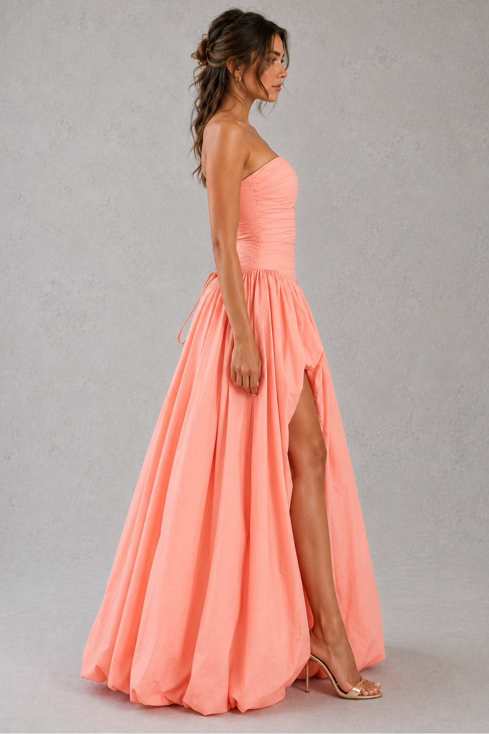 Woman wearing a coral pink strapless gown with a slit on a gray background