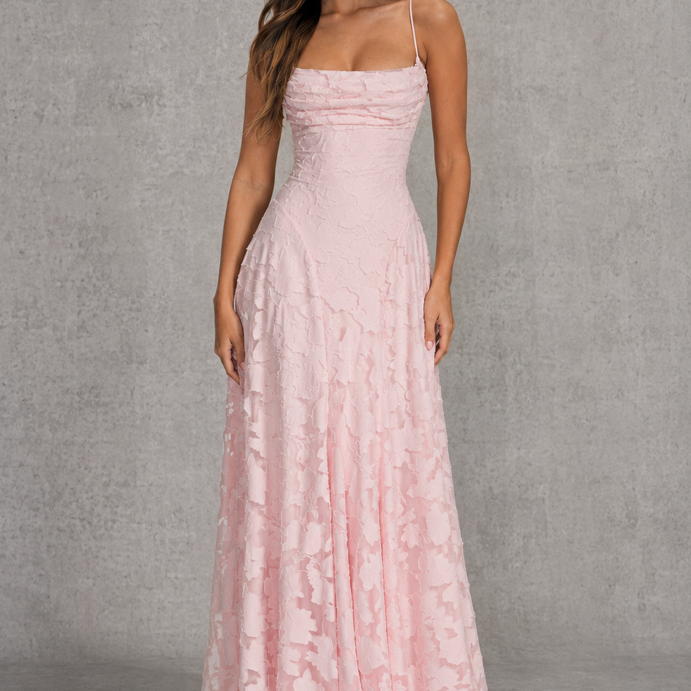 A woman modeling a floor-length light pink gown with thin straps, a fitted ruched bodice, and a flared skirt, standing against a textured gray studio backdrop.
