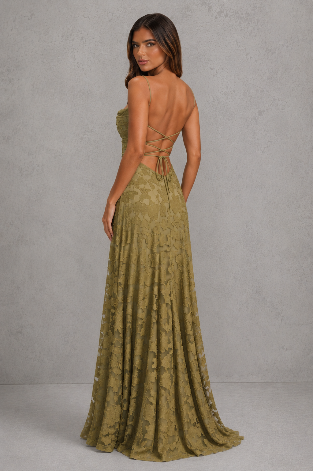 A woman modeling a floor-length olive green lace gown from the back, showing a strapless bodice, open back with tie details, and a full skirt against a textured gray studio backdrop.