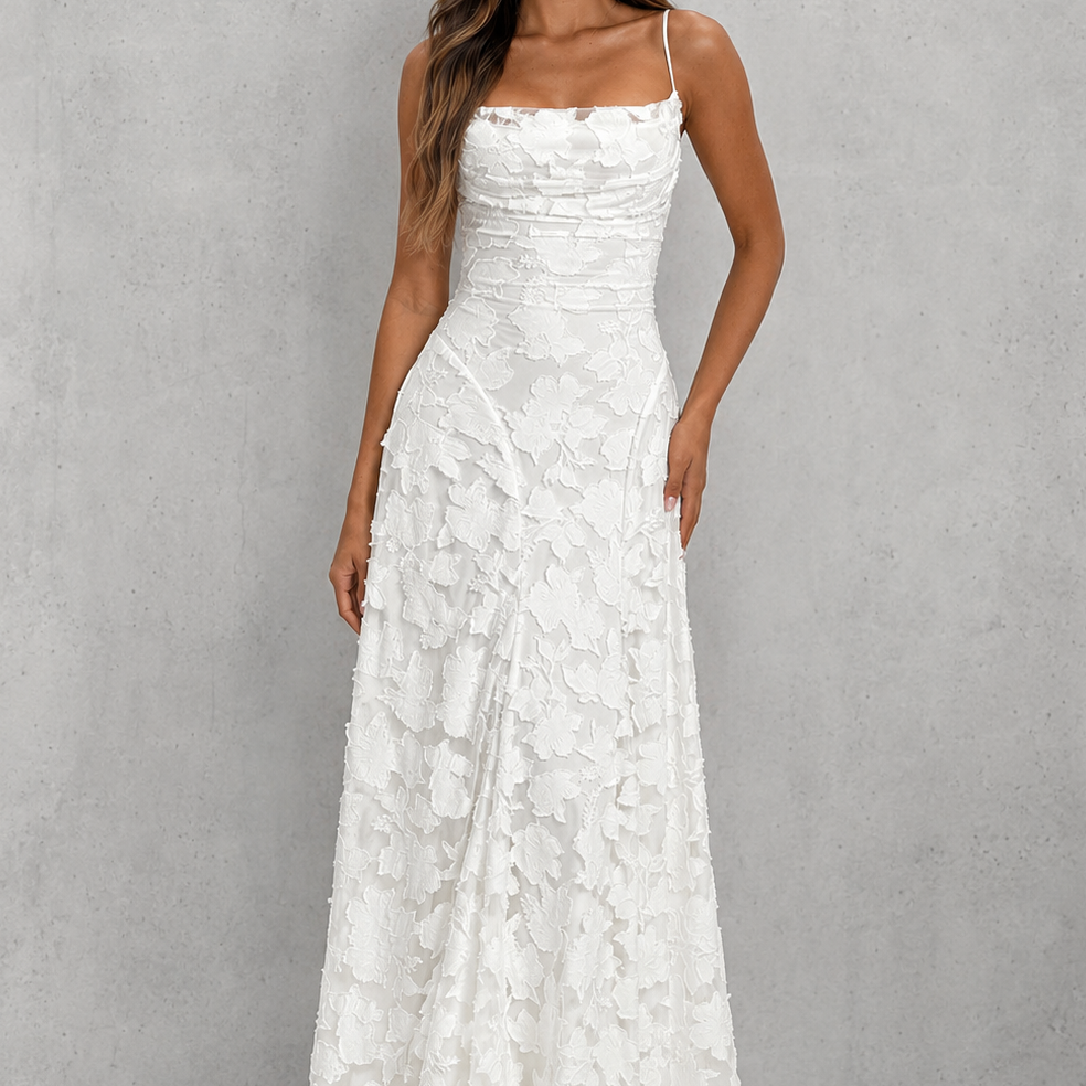 A woman modeling a floor-length white lace dress with thin straps and a fitted bodice, standing against a textured light gray studio backdrop.