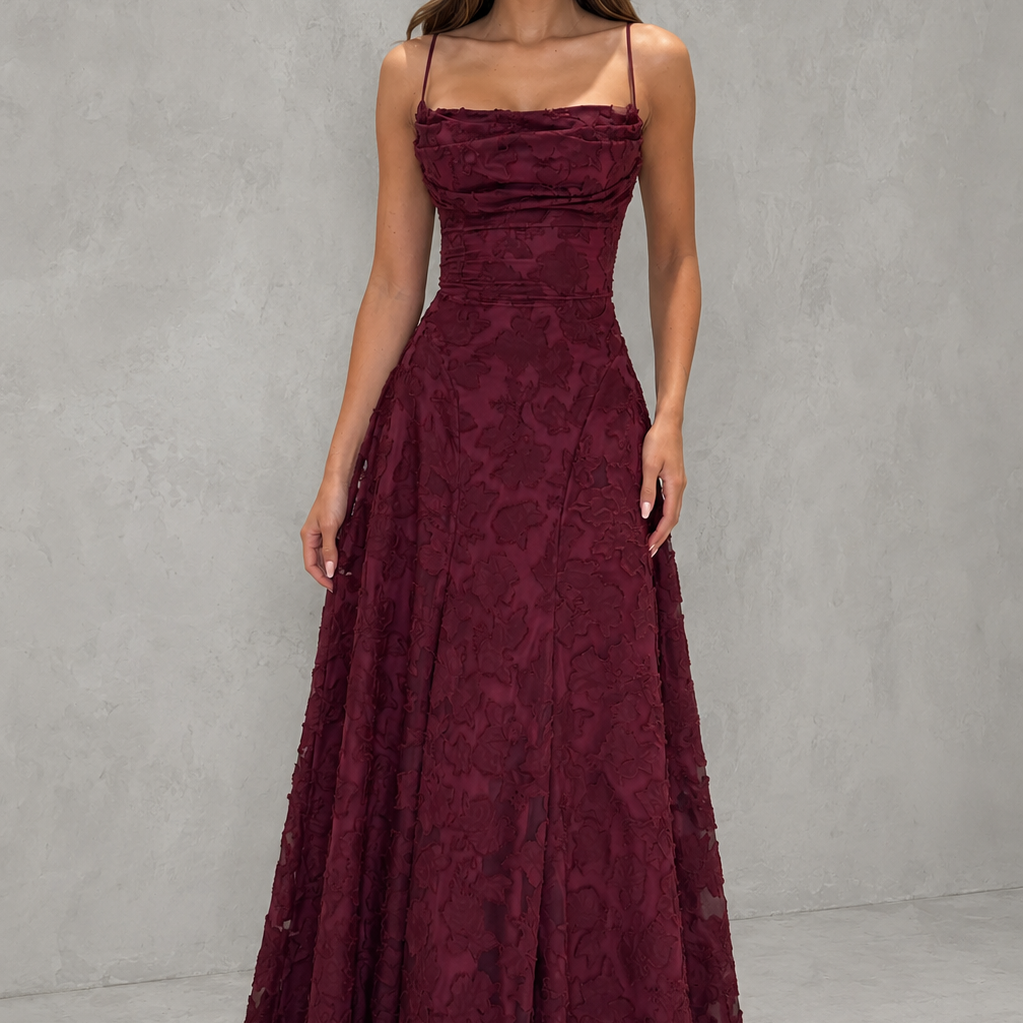 A woman modeling a floor-length burgundy lace gown with thin spaghetti straps, a structured sweetheart neckline, and a full A-line skirt, set against a textured gray studio backdrop.