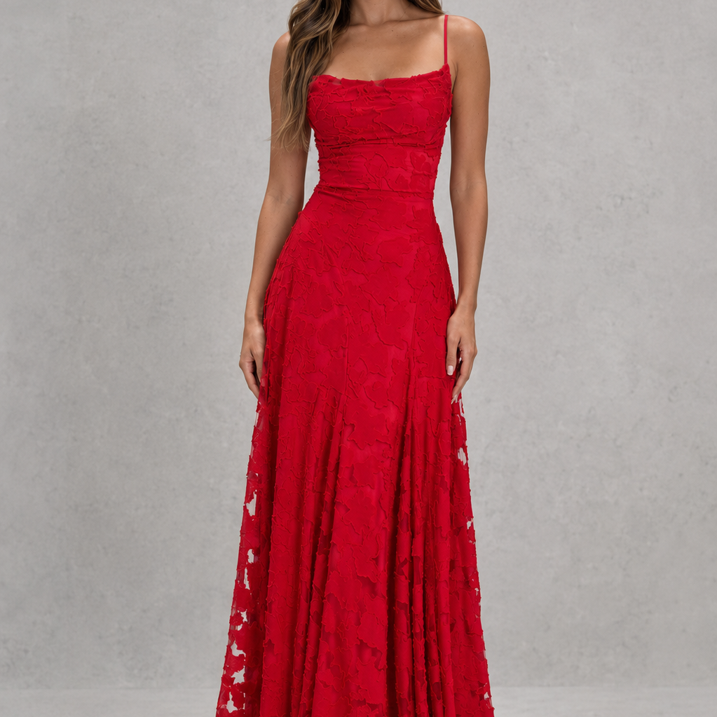 A woman modeling a floor-length red evening gown with thin straps, a fitted waist, and lace-up side details, photographed against a textured gray studio backdrop.