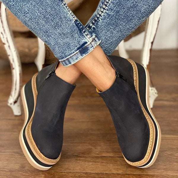Black slip-on shoes with beige soles worn by a person sitting on a wooden floor.