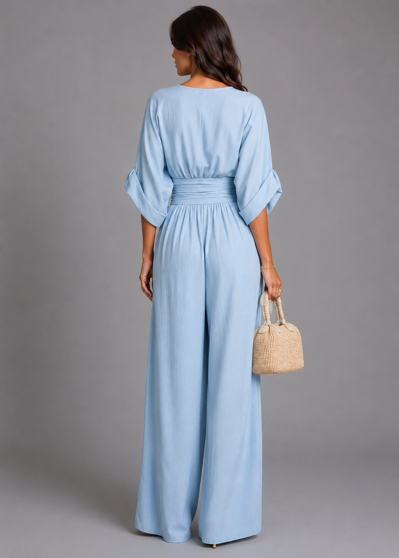 Woman wearing a light blue jumpsuit holding a woven bag against a gray background