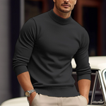 Man wearing a black turtleneck sweater and beige pants, standing in front of a blurred background.