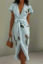 Woman wearing a light blue dress with floral patterns and holding a brown handbag against a plain background