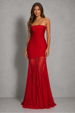 A woman modeling a strapless red gown with a fitted bodice, sheer mesh overlay at the upper legs, and a flowing skirt, photographed against a gray studio backdrop.